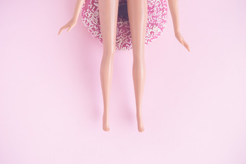 Flat lay of doll sitting on donut against pastel pink background.
