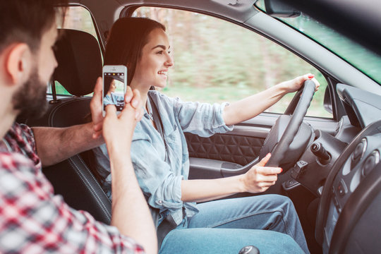 Young man is taking picture of girl driving car. He is doing that using phone. Girl is holding rudder with her hands and looking straight forward. She is smiling.