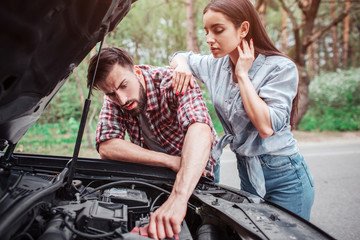 Smart and serious guy is fixing car. Girl is standing besides him and looking to the process. She is leaning on her boyfriend.