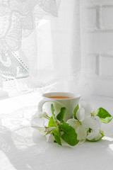 White cup with tea and centuries of blossoming apple trees in the summer morning breakfast at the window with a white lacy curtain in a rustic style with a copy of the space