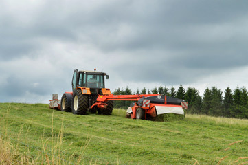 An agricultural tractor with a mower cutting long grass in a field.