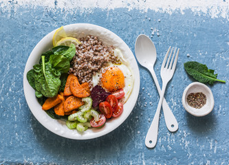 Full bowl healthy food - buckwheat, baked sweet potatoes, spinach, egg, beetroot, celery, tomatoes. Delicious diet lunch on blue background, top view