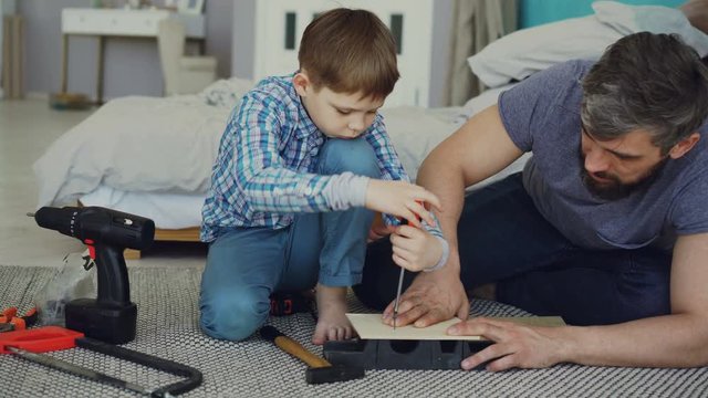 Curious child is driving screw in piece of wood with screwdriver and his dad is holding wooden sheet and teaching him basics of woodwork. United family concept.
