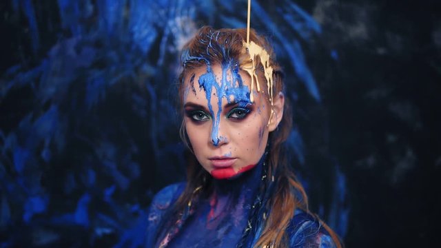 Front view of a beautiful lady in blue paint, pouring the yellow paint from the top down on her head and hair