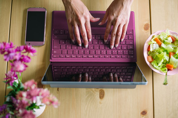 Close up of female hands typing on laptop. Working at home with modern technology concept. Convertible tablet, smartphone and healthy green salad.