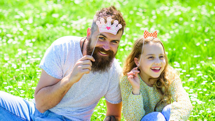 Fototapeta premium Family spend leisure outdoors, play girlish games. Child and father posing with crown and bow photo booth attributes. Fatherhood concept. Dad and daughter sits on grass at grassplot, green background.
