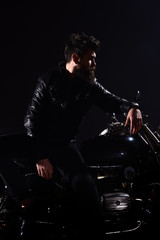 Obraz na płótnie Canvas Man with beard, biker in leather jacket lean on motor bike in darkness, black background. Brutality and masculine concept. Macho, brutal biker in leather jacket stand near motorcycle at night time.