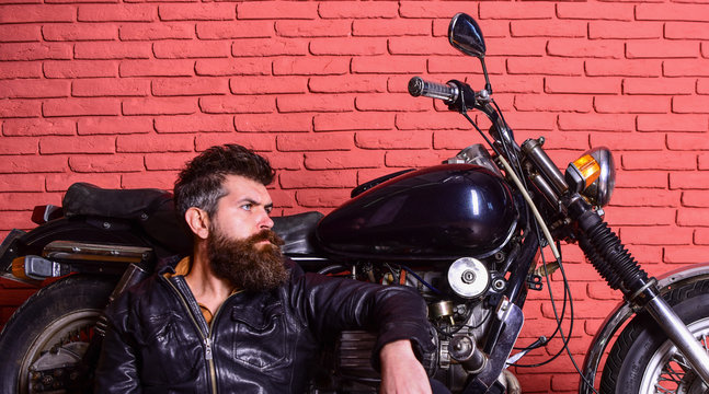 Man with beard, biker in leather jacket near motor bike in garage, brick wall background. Hipster, brutal biker on pensive face in leather jacket sits, leans on motorcycle. Bikers lifestyle concept.