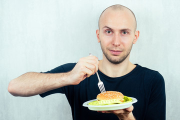 portrait of a young handsome bald man with a measuring tape eating a high-calorie big burger on a plate with a fork. concept of diet, unhealthy and healthy eating