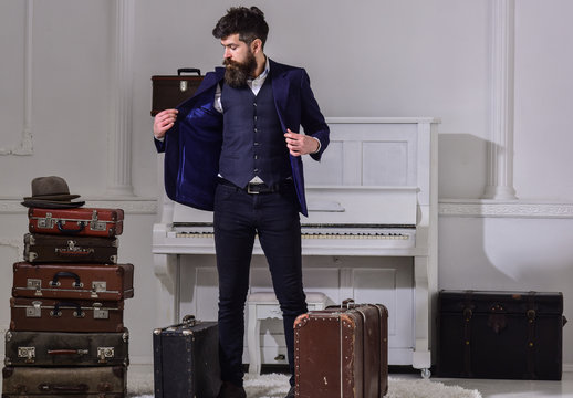 Hotel service concept. Man, traveller with beard and mustache with luggage, luxury white interior background. Macho elegant on strict face stands near pile of vintage suitcase, arrived to hotel.