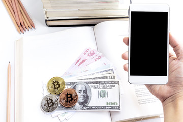 A woman's hand hold a smartphone with Banknote and bitcoin on the books open with pile textbooks and wooden pencils on the white table background. Money for education and business concept.