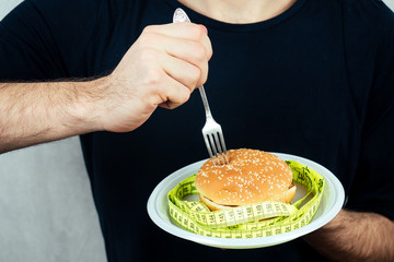 close-up hand of a man with a measuring tape eating a high-calorie big burger on a plate with a fork. concept of diet, unhealthy and healthy eating