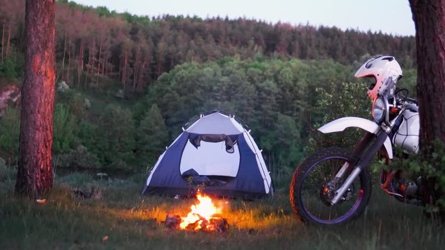 the motorcyclist man sits in a tourist tent in night and uses a smartphone, in the background a forest and a off road enduro motorcycle, camp travel, travel concept and active lifestyle, camp fire