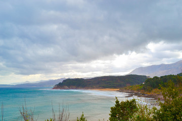 Obraz premium View of a beach in front of cliffs and hills, with green mountains and cloudy sky