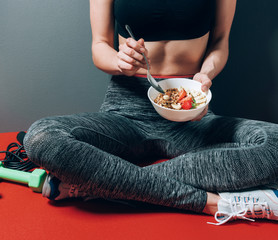 girl in sports clothes sitting near dumbbells and skipping rope with a plate of healthy breakfast...