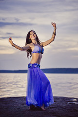 Fototapeta na wymiar Dancer of bellydance in a blue suit on the beach, against the background of the water. Beautiful nature at sunset. Belly dance. Girl with exotic appearance. Oriental beauty.
