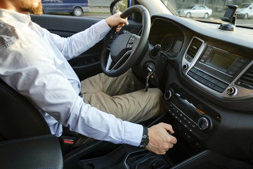 Close up of young man driving expensive car, seat belt strapped, hands w/ watch on steering wheel & shifting transmission gears, keys in ignition. Businessman rides his vehicle. Side view, background.