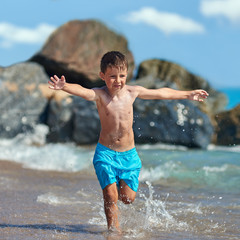 Cute Caucasian boy is running in the water along the sea shore against big boulders. His arms are wide open.