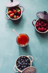 Top view of berries inside ceramic colored cocotte, blueberries, strawberries, raspberries, flat lay, blue table, wth a glass full of juice and spilling. Detox and healthy food concept.
