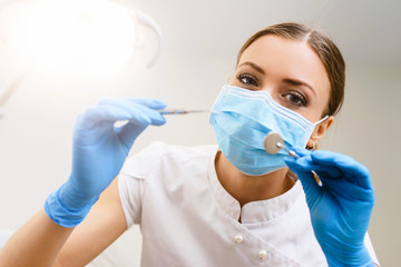 Young female friendly dentist in umiform standing in her office and looking at camera and smiling. Close up potrait of the doctor woman at white background
