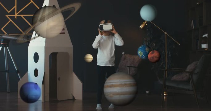 Little girl wearing VR virtual reality glasses standing near cardboard rocket, studying a model of a space solar system. 4K UHD 