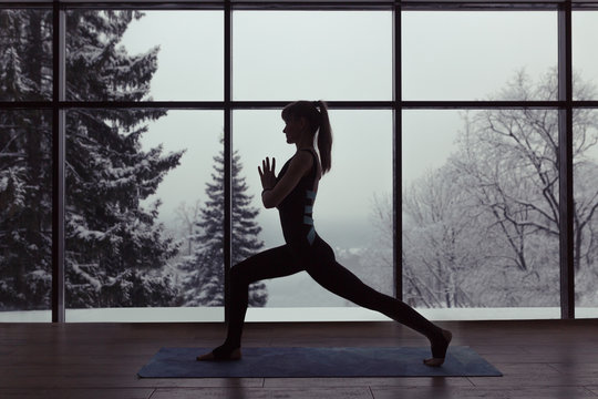 A Silhouette Of A Woman Doing Yoga On Background Of Windows With Beautiful Winter Landscape With Trees In The Snow And Sunlight, Young Yogi Girl In Warrior's Yoga Position