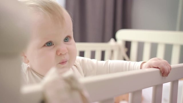 Sad baby standing in bed. Unhappy child holding on bed. Little baby emotion. Sad child standind in white cot. Toddler girl learn to stand in crib