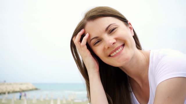 Portrait of happy smiling young woman sitting alone on beach. Wind from sea blowing female hair in slow motion. Vacation during off-season on Mediterranean Sea