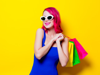 Young pink hair girl in blue dress with colored shopping bags. Portrait isolated on yellow background
