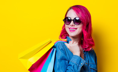 Young pink hair girl in sunglasses and blue shirt holding a colored shopping bags. Portrait on isolated yellow background