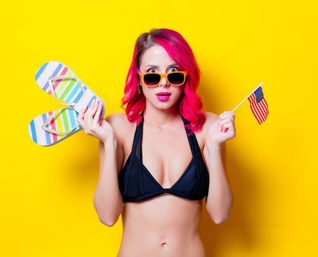 Young pink hair girl in bikini and orange glasses with flip flops and american flag. Portrait isolated on yellow background