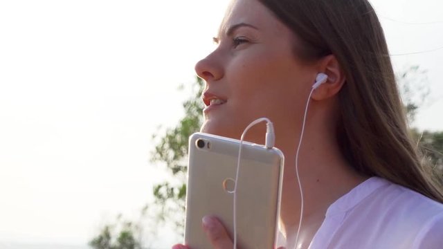 Close up of young happy woman in white shirt using mobile in green park. Female listening music via app on cellphone and singing along. Wind blowing her hair in slow motion