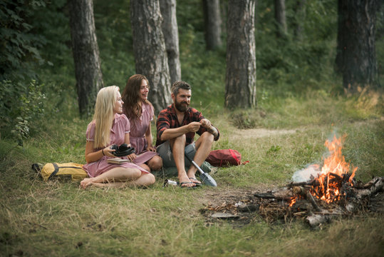 Happy friends enjoying picnic in woods. Bearded man and two sisters in retro dresses sitting next to campfire and cooking food outdoors
