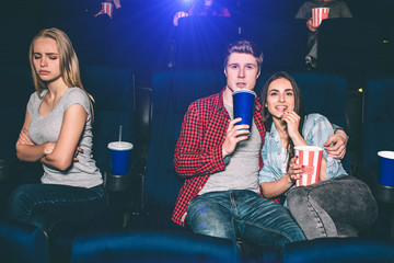 Beautiful couple is sitting together and watching movie. Brunette girl is eating popcorn and guy is drinking coke. She is offended of being alone.