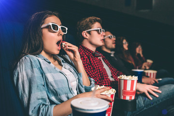 Amazed and surprised people are sitting in one row and watching movie. Blonde girl is eating popcorn with amazement on her face because of movie. The other people are amazed as well.