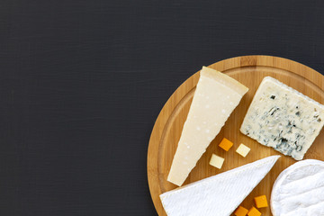 Various types of cheese on round wooden board, top view. Dark background. Copy space. Flat lay. From above.