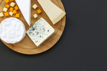 Various types of cheese on round wooden board, top view. Dark background. Copy space. Flat lay. From above.