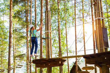 Teenage girl goes on hinged trail in extreme rope Park in summer forest. High-altitude climbing training of child on adventure track, equipped with safety straps and protective helmet. Estonian summer