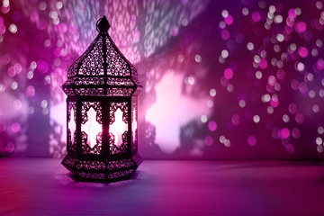 Ornamental Arabic lantern with burning candle glowing at night and glittering colorful bokeh lights. Festive greeting card, invitation for Muslim holy month Ramadan Kareem. Blurred party background