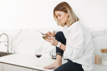 Portrait of young lady sitting on kitchen counter with cellphone and credit card in hands and glass of red wine near at home isolated