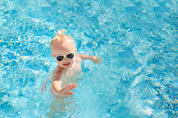 Fototapeta na wymiar Cute little baby in sunglasses swimming in the pool and waving his hand. Place for text. The view from the top.