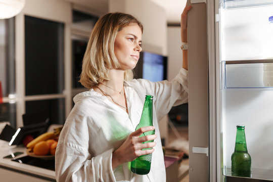 Portrait Of Young Lady Standing On Kitchen At Night And Thoughtfully Looking In Open Fridge While Holding Beer In Hand At Home Isolated