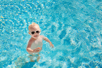 Cute little baby with sunglasses splashing happily in the pool with clear blue water in the summer. Top view. Copy space.