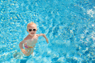 Fototapeta na wymiar Little blonde girl swims in the pool with blue water. The view from the top. Place for text.