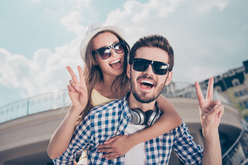 Love story! Attractive joyful cheerful tourists having journey trip meeting outdoor in sunny day showing v-signs, handsome man with bristle and headphones carrying on back his beautiful woman in cap