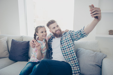 Cute girl with bearded stylish father in casual outfit checkered shirts sitting on couch in house embracing shooting self portrait on front camera showing peace symbol holding cell smart phone in hand