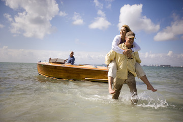 Smiling mature man piggybacking his wife from a boat to the shore on a beach.