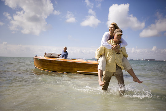 Smiling mature man piggybacking his wife from a boat to the shore on a beach.
