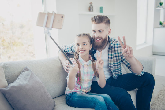 Portrait of cheerful friendly daddy and cute schoolgirl sitting indoor with pigtails using stick smart phone shooting selfie on front camera having video call showing two fingers hi sign