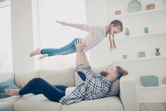 Side View Portrait Of Family With One Parent, Strong Stylish Father Lying On Couch Lifting Up His Daughter Making Plane Holding Hands To The Side Enjoying Time Together Indoor In Modern House